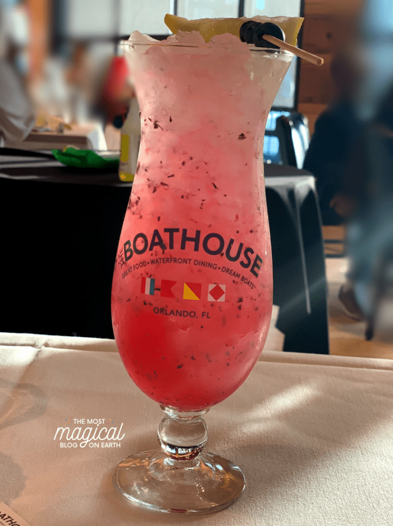 bell shaped fluted glass with words boathouse printed on the front in blue text, filled with ice and pink liquid and a pineapple on top